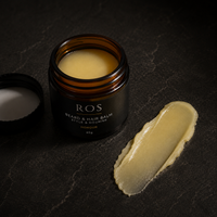 An open amber glass jar of ROS Beard & Hair Balm with its black lid placed beside it, alongside a smear of the pale yellow balm on a dark textured surface.