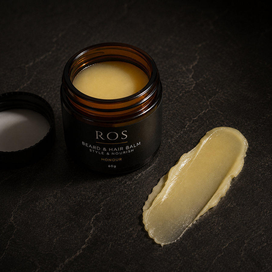 An open amber glass jar of ROS Beard & Hair Balm with its black lid placed beside it, alongside a smear of the pale yellow balm on a dark textured surface.