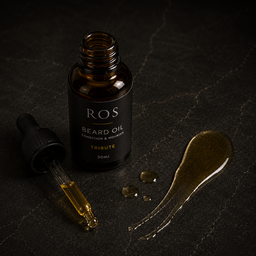 A 30ml bottle of ROS Tribute Beard Oil with its dropper beside a smear of golden beard oil on a dark textured background.