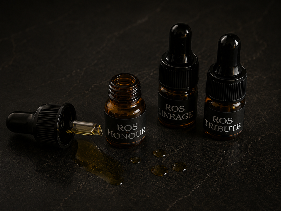 Three amber glass dropper bottles with beard oil scent samples discovery kit black caps and black labels reading ROS HONOUR, ROS LINEAGE, and ROS TRIBUTE, arranged on a dark stone surface with one bottle lying on its side and a few drops of golden liquid spilled in front.